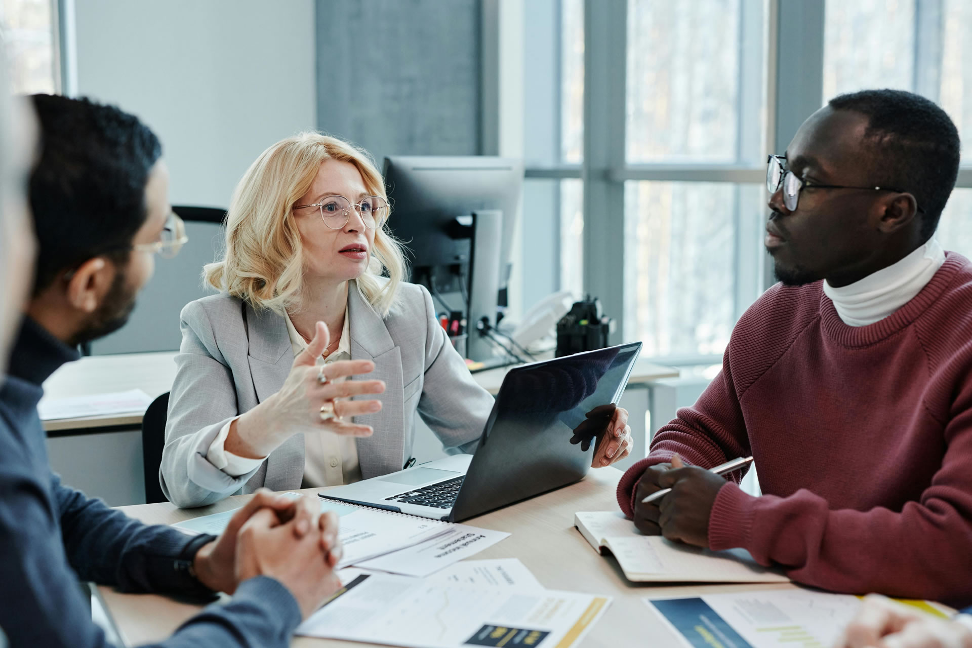 A group of people in a Accounting meeting, with a woman gesturing while talking. Papers and a laptop are on the table.