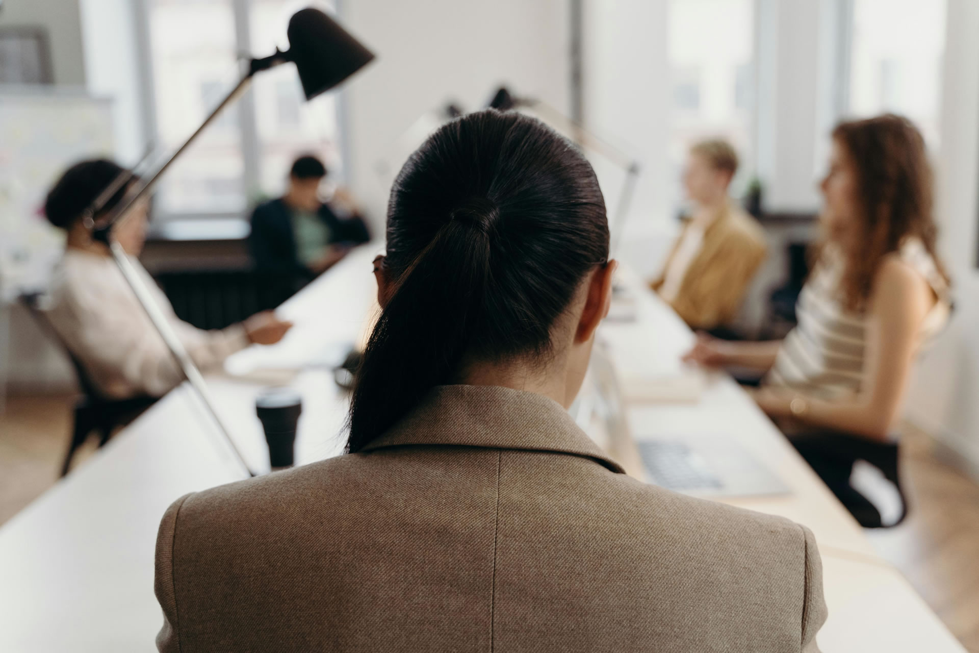 A person with a ponytail sits at a desk in an office, facing away. Three other people are working at their desks in the background.